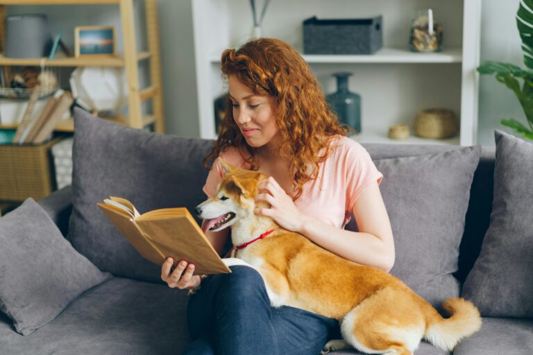 Redheaded woman reads a book with her Shiba Inu dog on a comfy sofa at home.